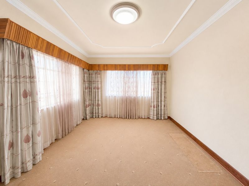 A spacious, empty living room featuring neutral walls, carpeted flooring, decorative ceiling cornices, and timber pelmets over large windows.