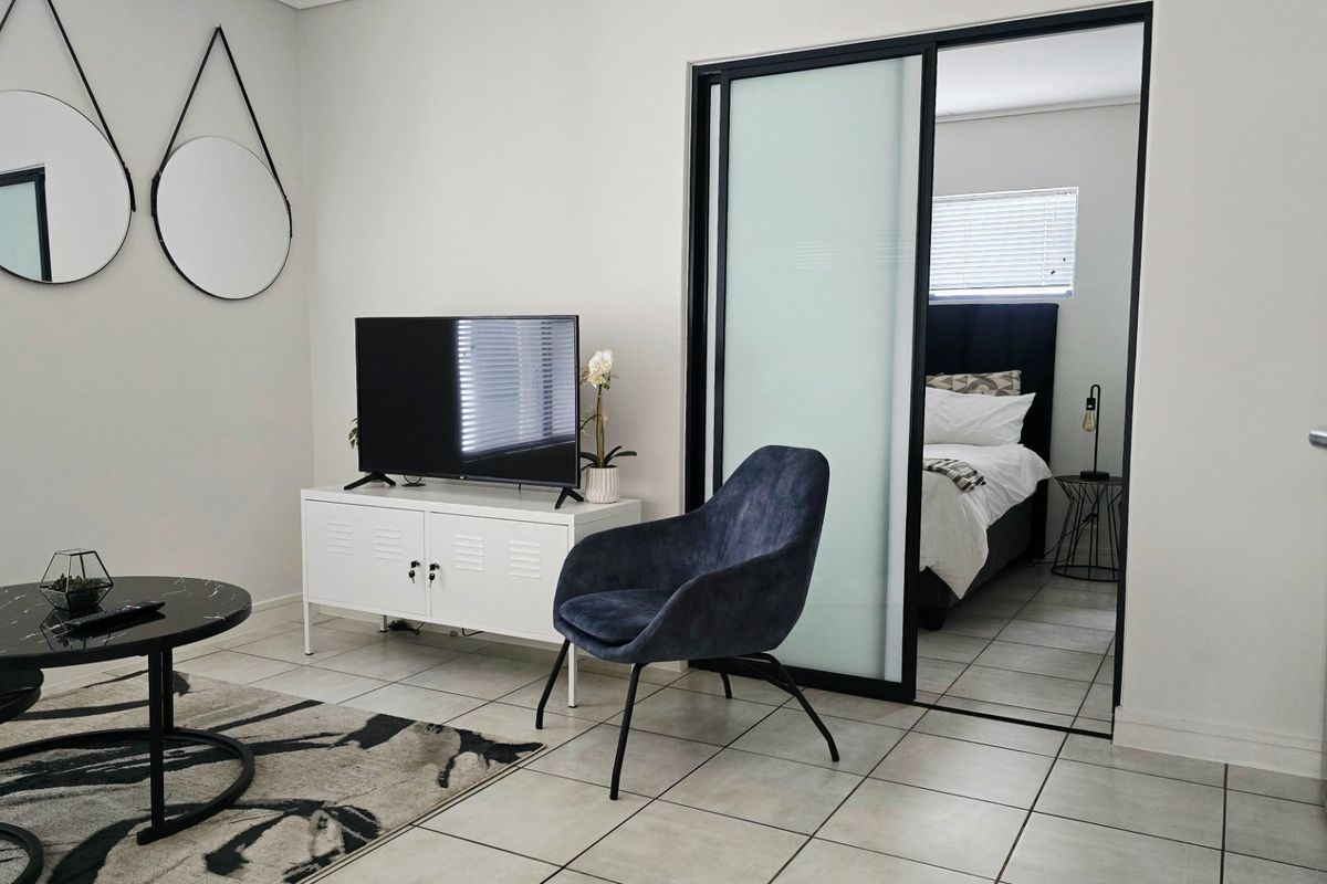 A modern living area featuring neutral tiled flooring and a sleek black-framed sliding glass door leading into the adjacent bedroom.