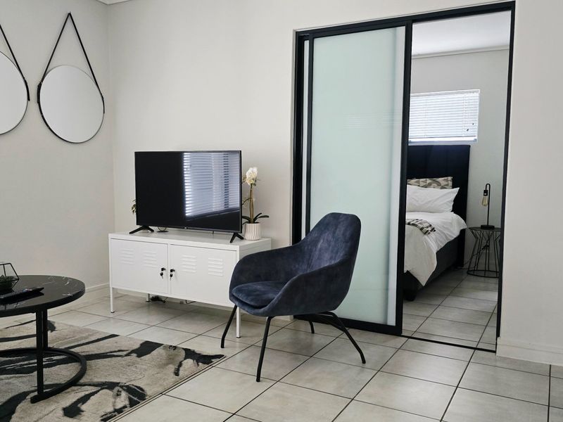 A modern living area featuring neutral tiled flooring and a sleek black-framed sliding glass door leading into the adjacent bedroom.