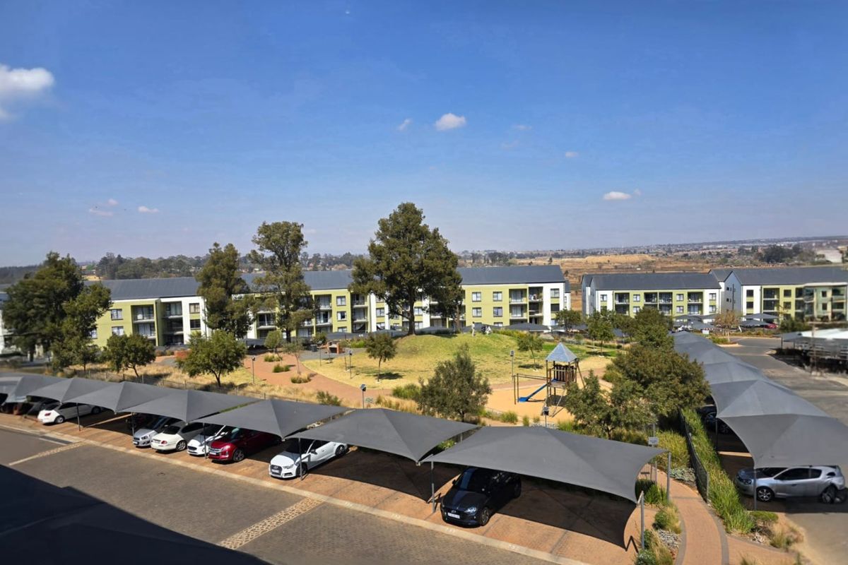 Aerial view of an apartment complex featuring communal landscaped gardens, a children's playground, and covered shade cloth parking bays.