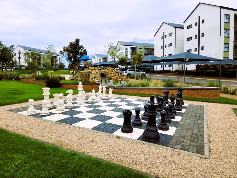 Outdoor communal area featuring a large paved chess board, landscaped gardens, and playground facilities within a well-maintained apartment complex.