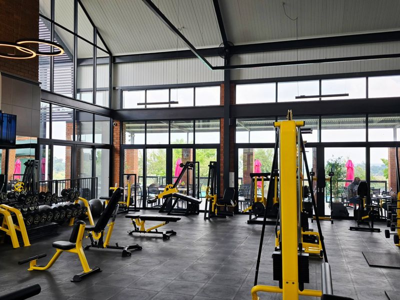 A modern, well-lit gym facility featuring high vaulted ceilings, exposed steel structural elements, and extensive floor-to-ceiling glass windows overlooking the outdoor area.