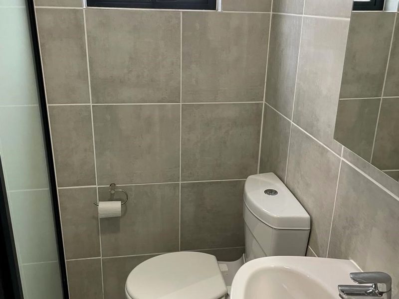 A modern, compact bathroom featuring neutral grey wall and floor tiles, a white toilet, a pedestal washbasin with a mixer tap, and a shower enclosure.