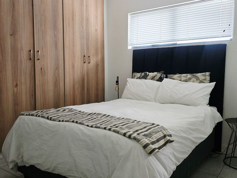 A well-maintained bedroom featuring modern wood-finish built-in cupboards, tiled flooring, and a window fitted with venetian blinds.