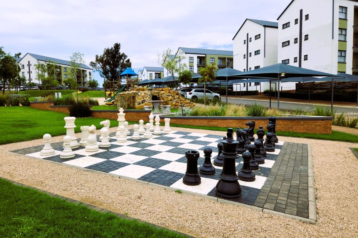A well-maintained communal garden area featuring a large outdoor chess set, paved pathways, and shade structures within an apartment complex.