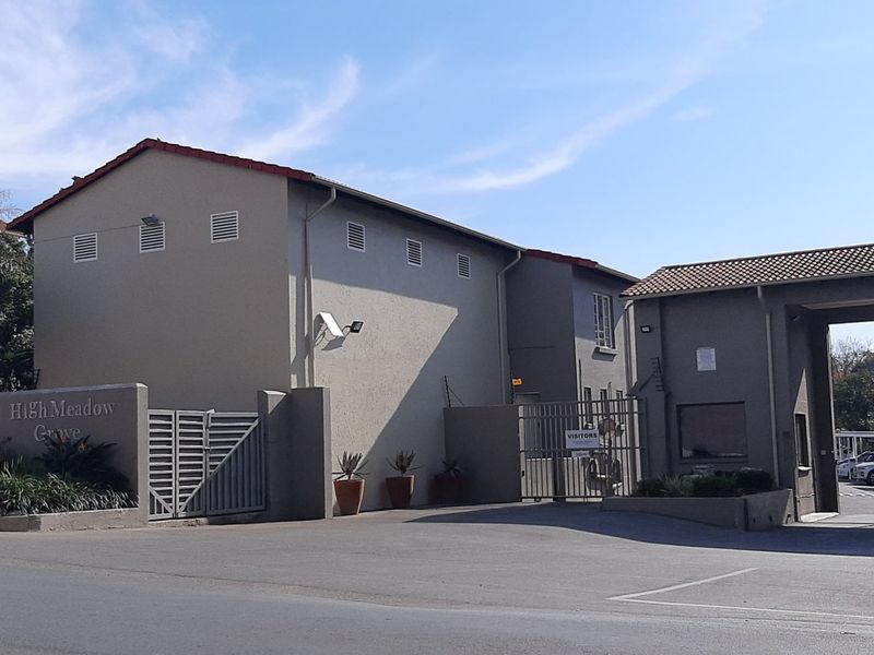 Front entrance view of High Meadow Grove apartment complex featuring a secure gated access point, paved driveway, and perimeter walling.
