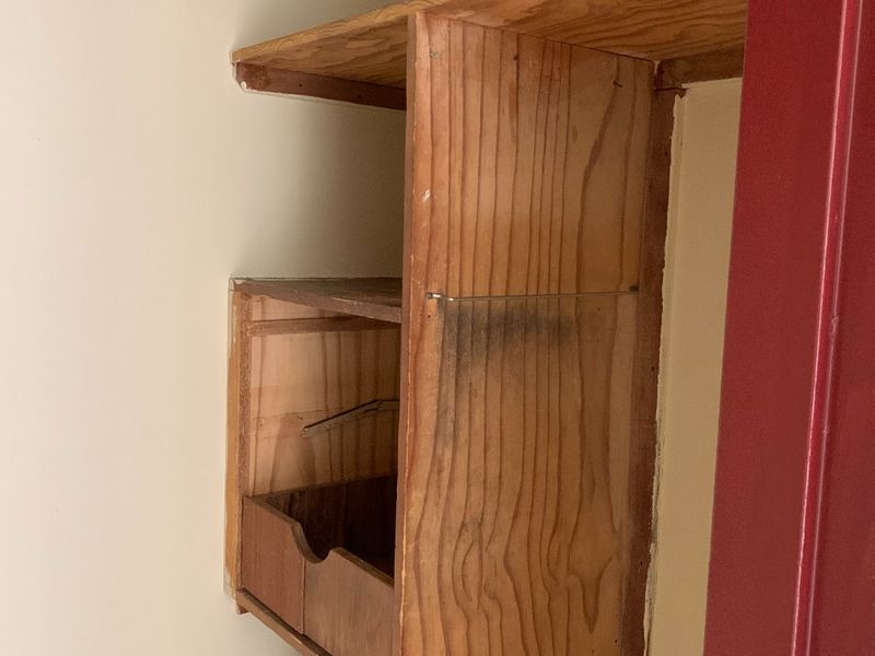A small utility or storage closet featuring dated, worn built-in wooden shelving and storage drawers in need of refurbishment.