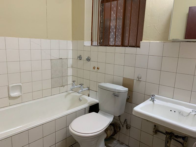 A dated bathroom featuring a standard bathtub, toilet, and wall-mounted basin with tiled walls, requiring significant renovation and cosmetic updates.