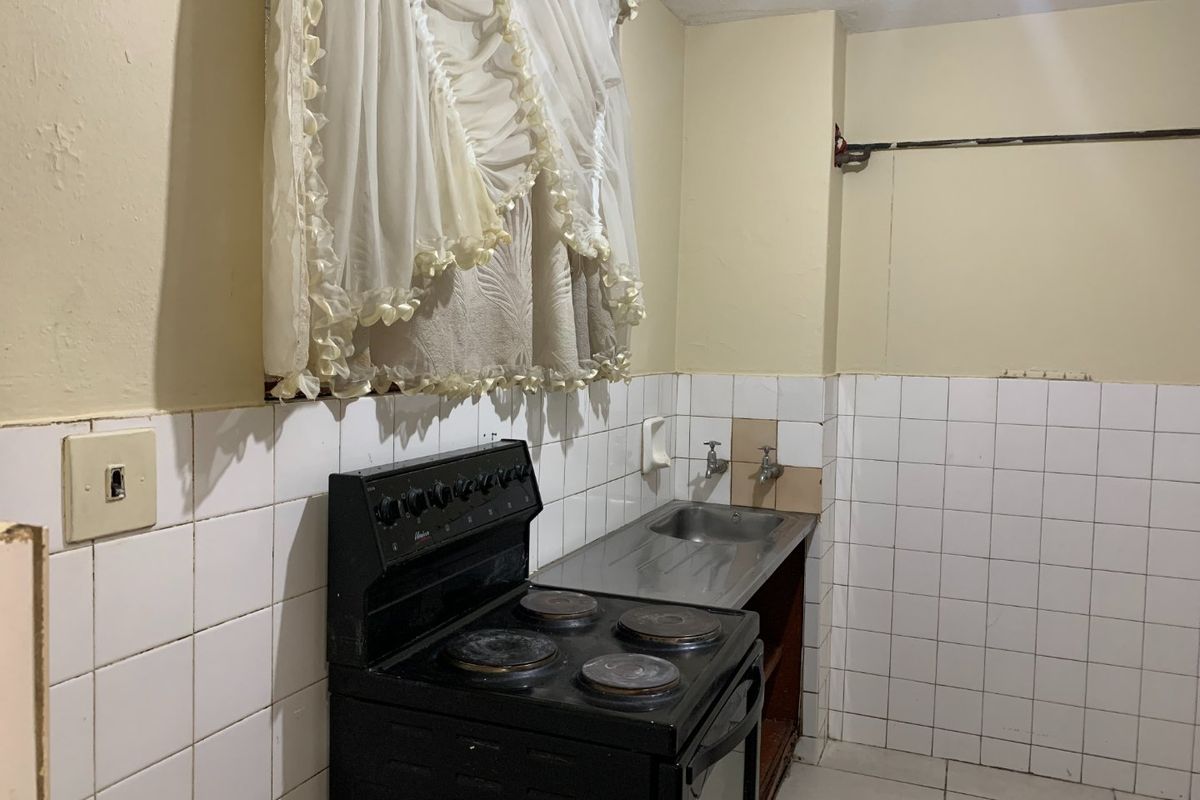 A compact kitchen area featuring a basic electric stove, stainless steel sink, and tiled splashback, requiring cosmetic updates to improve its overall condition.