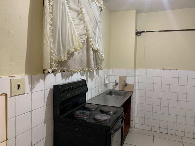 A compact kitchen area featuring a basic electric stove, stainless steel sink, and tiled splashback, requiring cosmetic updates to improve its overall condition.
