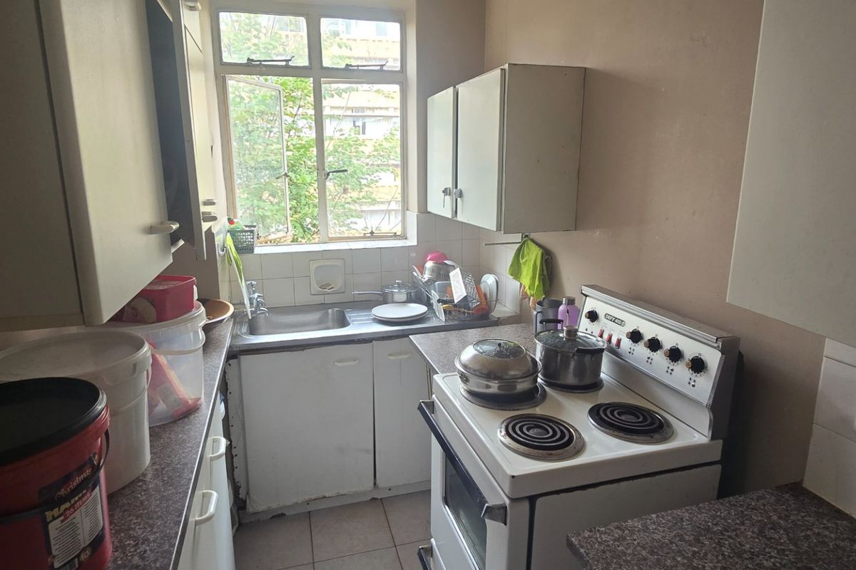 A compact, dated kitchen featuring original cabinetry, a freestanding electric stove, and a stainless steel sink with a window view.