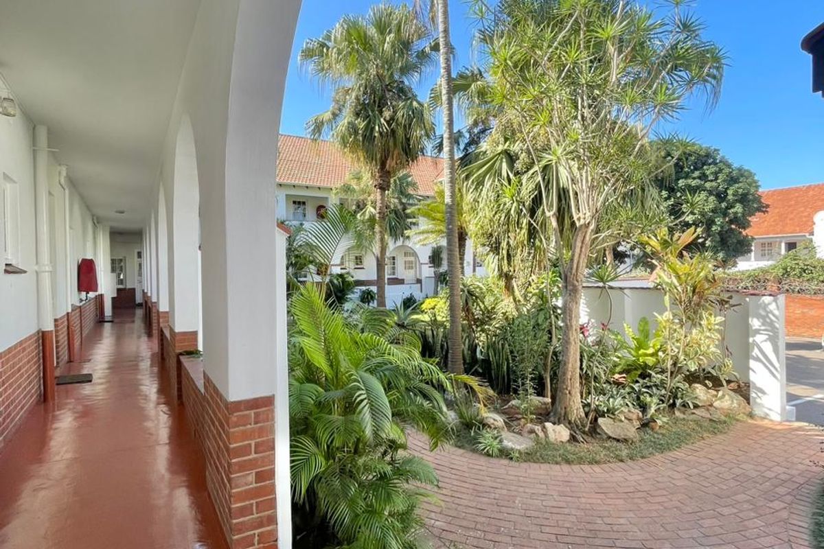 A well-maintained covered walkway with arched architectural features overlooking a landscaped courtyard with brick paving and mature palm trees.