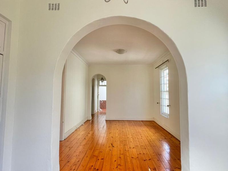 An empty living area featuring polished hardwood floors, a classic architectural archway, and a decorative chandelier, offering a bright and open living space.
