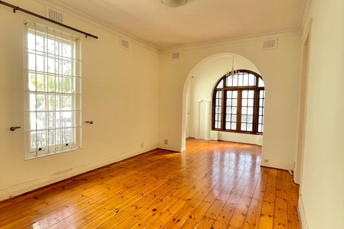 A bright, empty living room featuring polished timber flooring, an elegant archway, and large timber-framed windows with security bars.