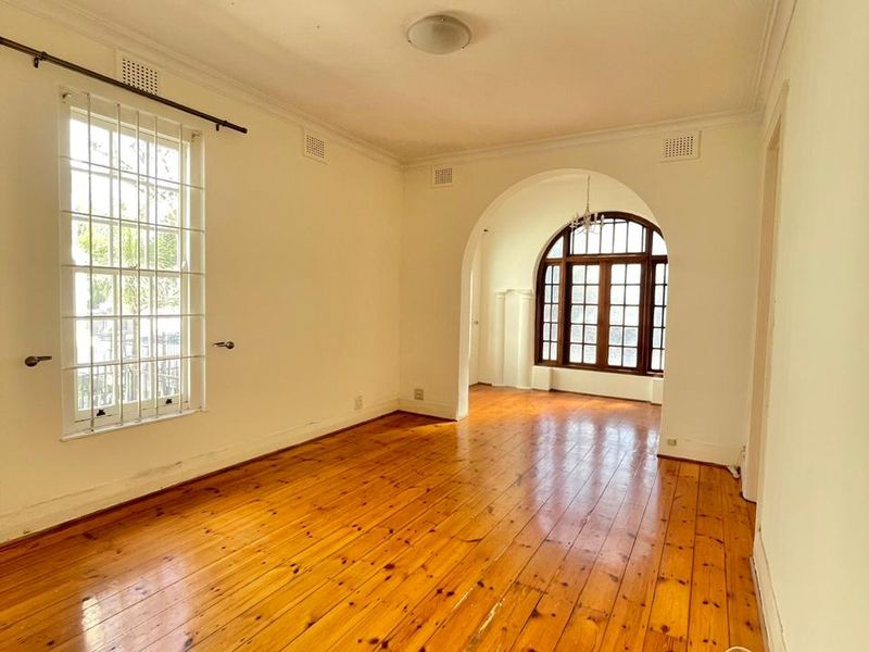A bright, empty living room featuring polished timber flooring, an elegant archway, and large timber-framed windows with security bars.