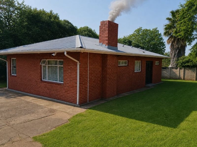 Rear view of a single-story red brick house featuring a prominent chimney, corrugated metal roof, and surrounding lawn area.