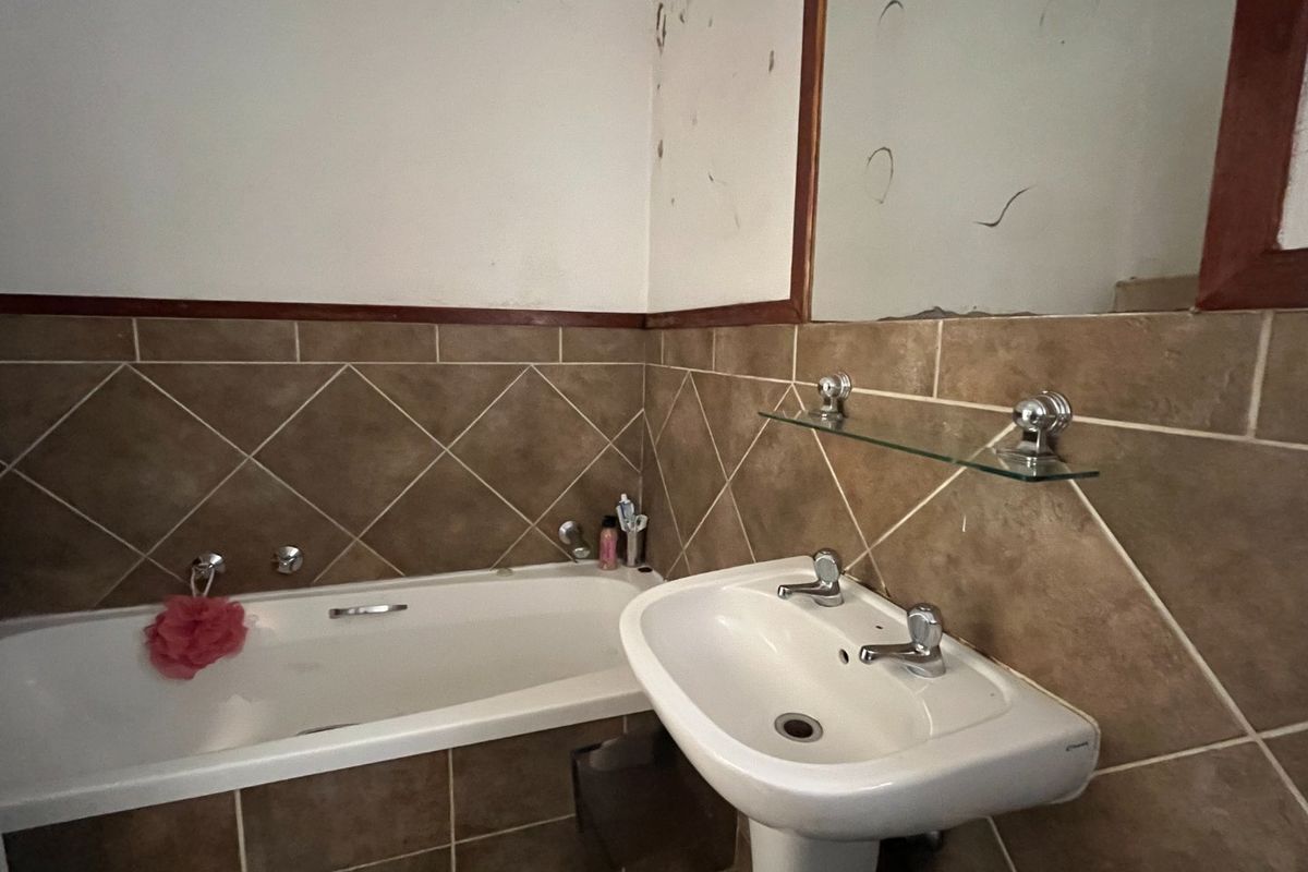 A dated bathroom featuring a pedestal basin, built-in bathtub, and brown ceramic wall tiling, requiring cosmetic updates and repairs to the wall and mirror.