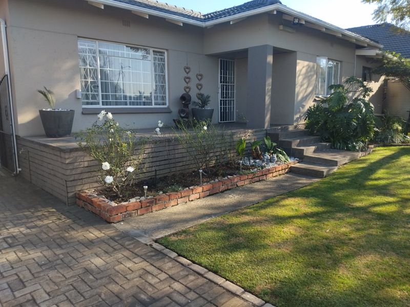 Front elevation of a single-story house featuring a paved driveway, brick retaining wall, landscaped garden beds, and a covered entrance with security gate.