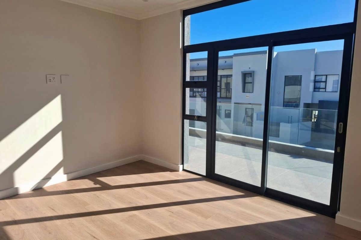 A bright, empty bedroom featuring modern laminate flooring and large sliding glass doors that open onto an outdoor balcony area.