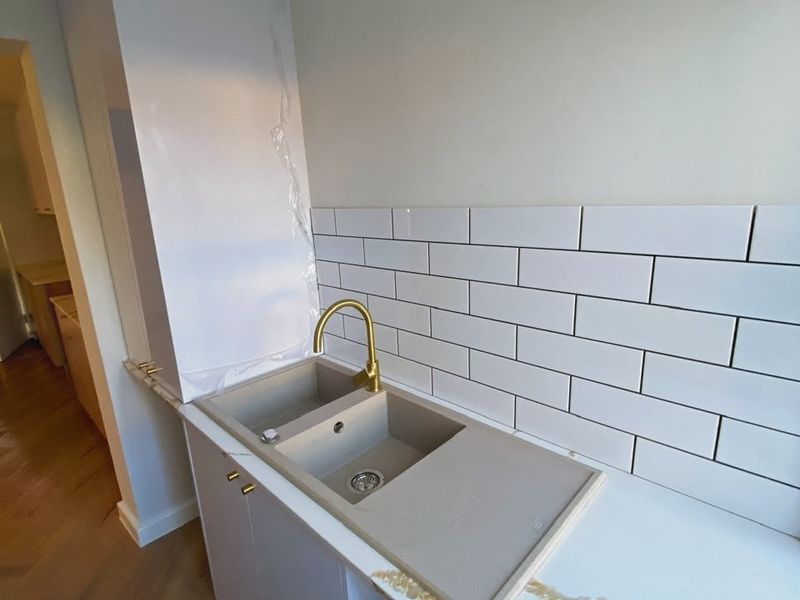 A modern kitchen area featuring a composite double sink, brass faucet, white subway tile backsplash, and elegant stone countertops with herringbone timber flooring.