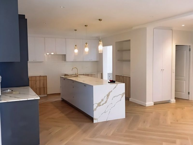 Modern open-plan kitchen featuring a large marble-look island, herringbone timber flooring, custom cabinetry, and elegant brass fixtures in a bright, contemporary space.