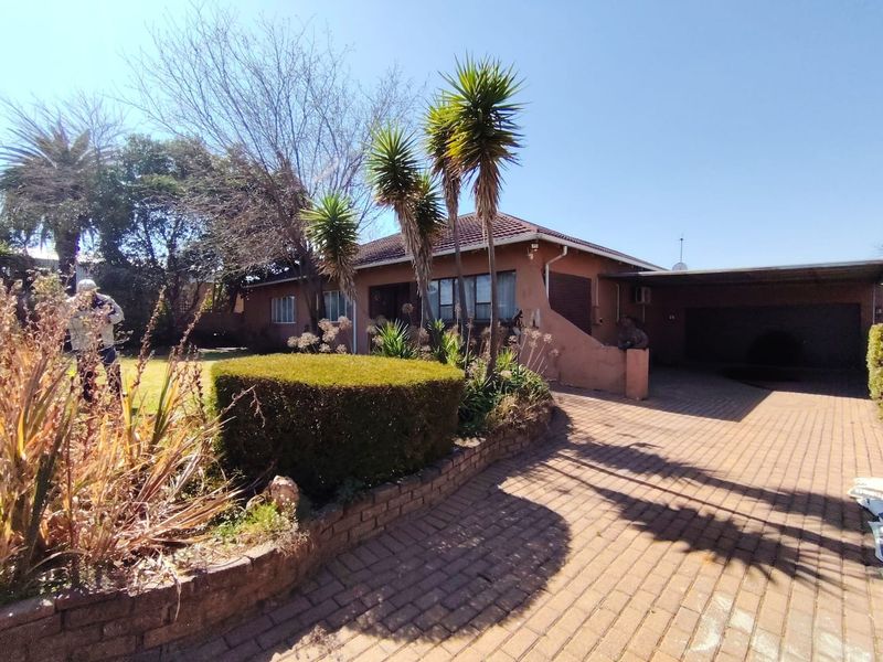 Front elevation of a single-story house featuring a paved driveway, attached carport, and established garden with a brick retaining wall.