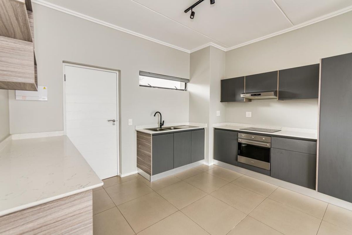 Modern kitchen featuring sleek dark cabinetry, stone countertops, integrated stainless steel oven, electric hob, and contemporary track lighting in an open-plan layout.