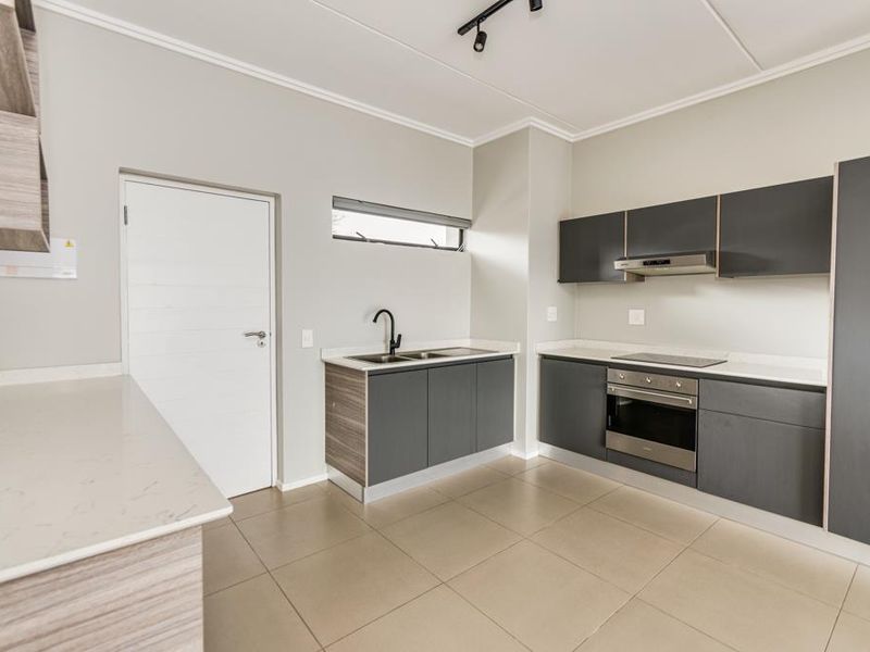 Modern kitchen featuring sleek dark cabinetry, stone countertops, integrated stainless steel oven, electric hob, and contemporary track lighting in an open-plan layout.