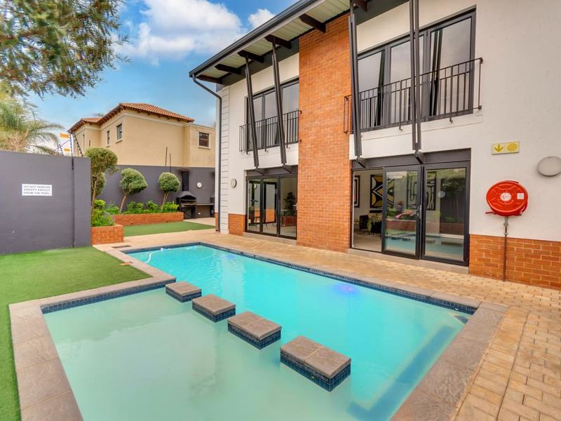 A well-maintained outdoor pool area featuring a swimming pool with stepping stones, paved patio, artificial lawn, and a modern brick and glass building facade.