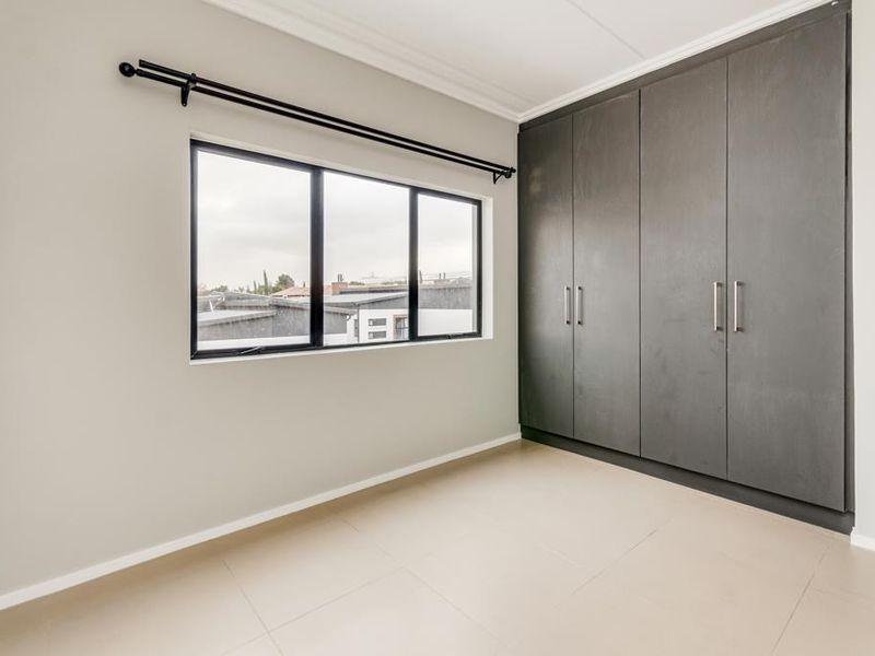 A bright, empty bedroom featuring neutral tiled flooring, modern dark-toned built-in cupboards, and a large window with a black frame.