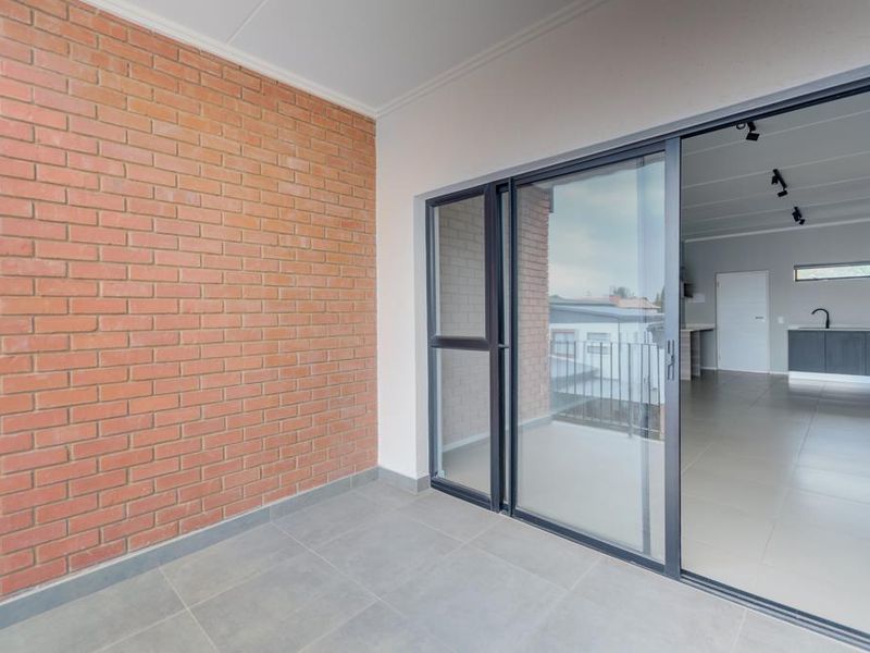 A clean, modern balcony featuring an exposed brick feature wall, tiled flooring, and large sliding glass doors leading into the apartment interior.