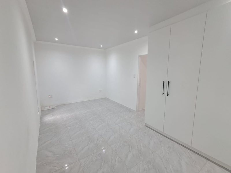 A bright, modern bedroom featuring neutral tiled flooring, built-in cupboards, and recessed downlighting, presented in a clean, vacant state.
