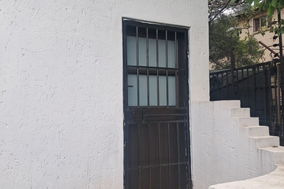 Exterior view of a rear building entrance featuring a security gate, textured plaster walls, and concrete steps leading to the doorway.