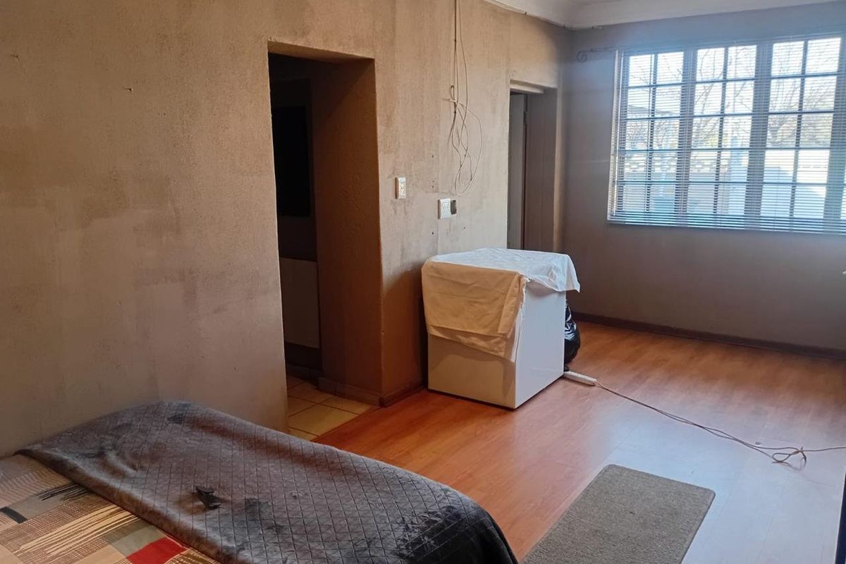 A bedroom featuring laminate flooring and a window with blinds, currently requiring wall finishing and electrical repairs to improve the overall condition.