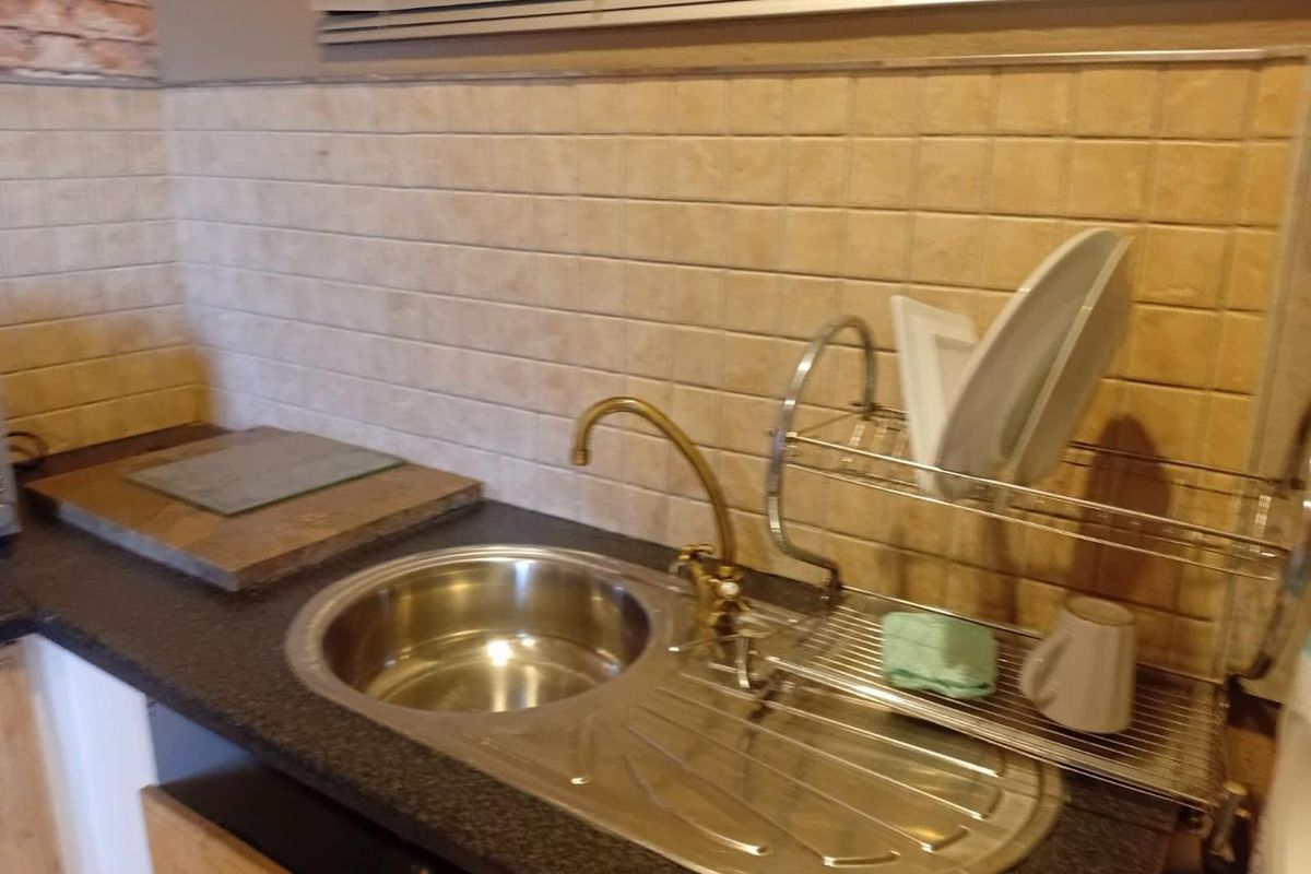 A functional kitchen area featuring a stainless steel sink with a brass faucet, dark laminate countertops, and a neutral tiled backsplash.