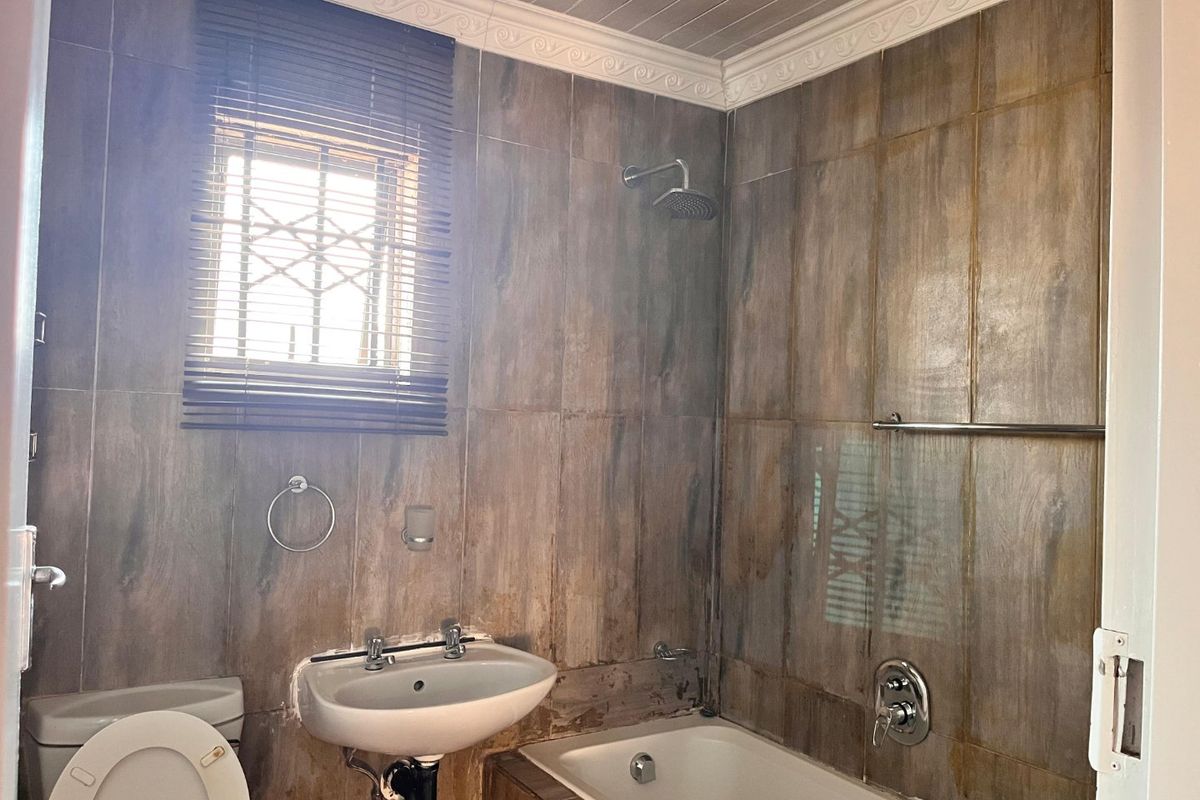 A dated bathroom featuring wood-look wall tiles, a built-in bathtub with shower, pedestal basin, and toilet, requiring cosmetic updates to modernize the space.