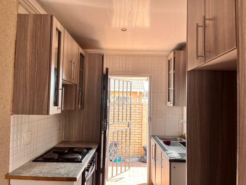 Compact galley-style kitchen featuring wood-look cabinetry, built-in electric hob and oven, tiled backsplash, and direct access to an exterior courtyard.