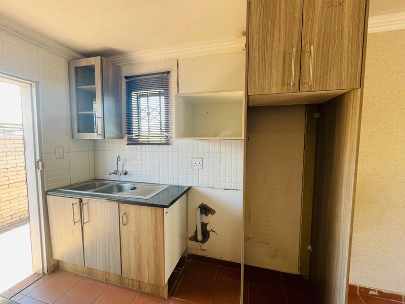 A compact kitchen featuring laminate cabinetry, a double sink, and tiled backsplash, currently requiring repairs to the wall and plumbing area.