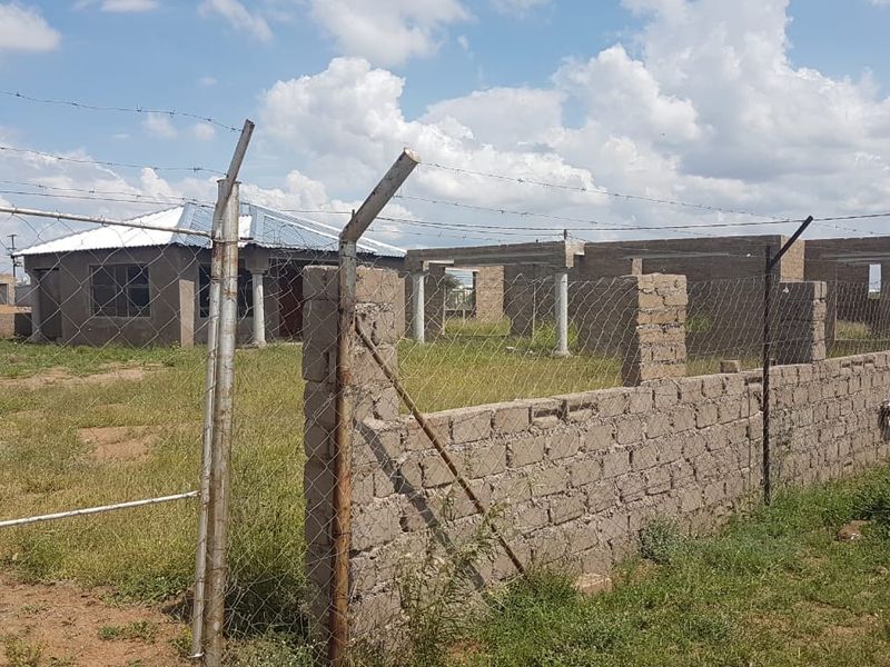 A view of a property under construction, featuring an unfinished building structure enclosed by a concrete block wall and barbed wire fencing.
