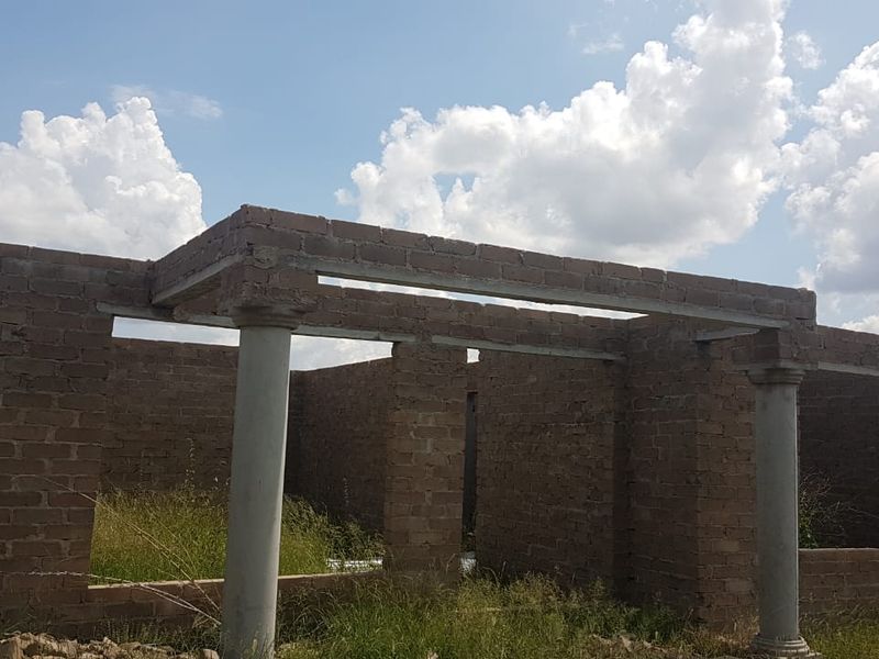 An unfinished residential structure showing exposed brick masonry walls and concrete support columns, currently in the early stages of construction.