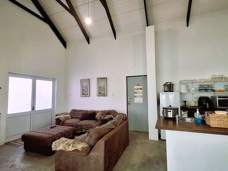 Open-plan living area featuring a high vaulted ceiling with exposed timber beams, integrated kitchen space with open shelving, and direct access to the outdoors.