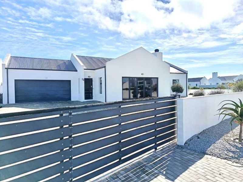 Modern single-story house featuring a white facade, corrugated metal roof, paved driveway, and a secure slatted metal gate entrance.