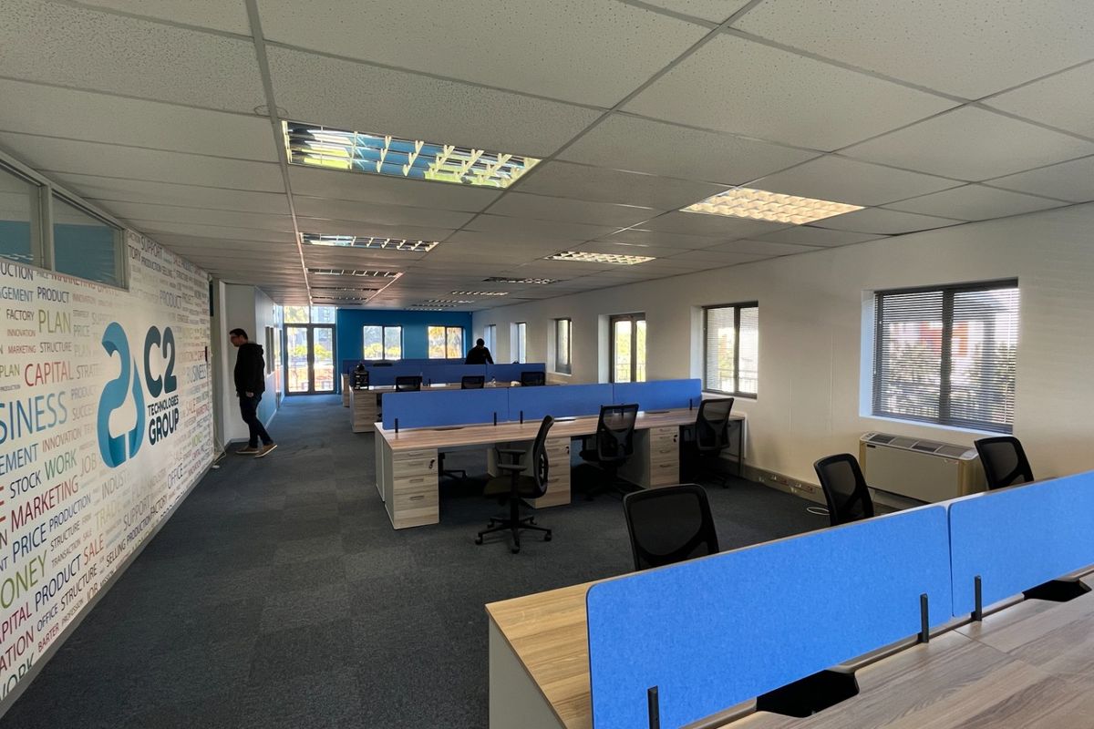 Open-plan commercial office space featuring a suspended ceiling, recessed lighting, carpeted flooring, and built-in HVAC units with large windows providing natural light.