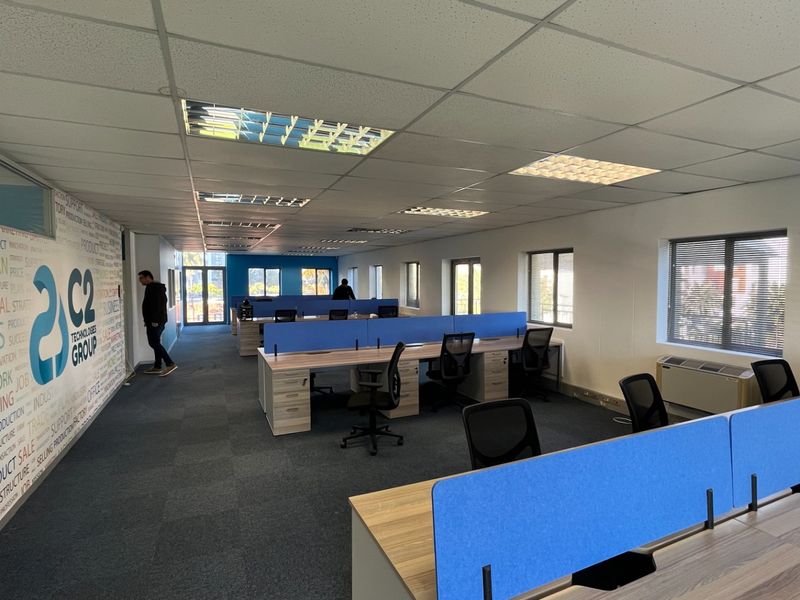 Open-plan commercial office space featuring a suspended ceiling, recessed lighting, carpeted flooring, and built-in HVAC units with large windows providing natural light.