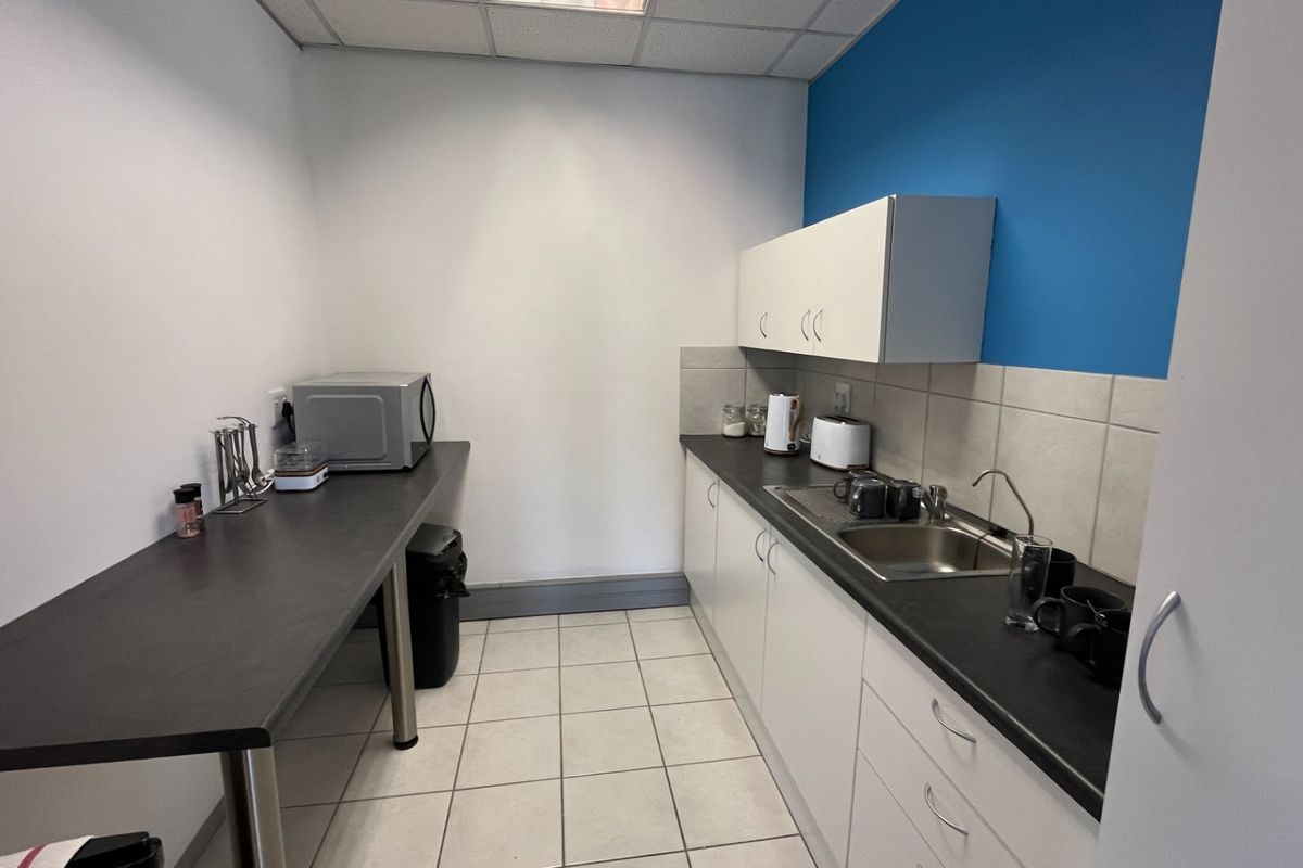 A functional commercial kitchenette featuring white cabinetry, dark laminate countertops, a stainless steel sink, and tiled flooring.