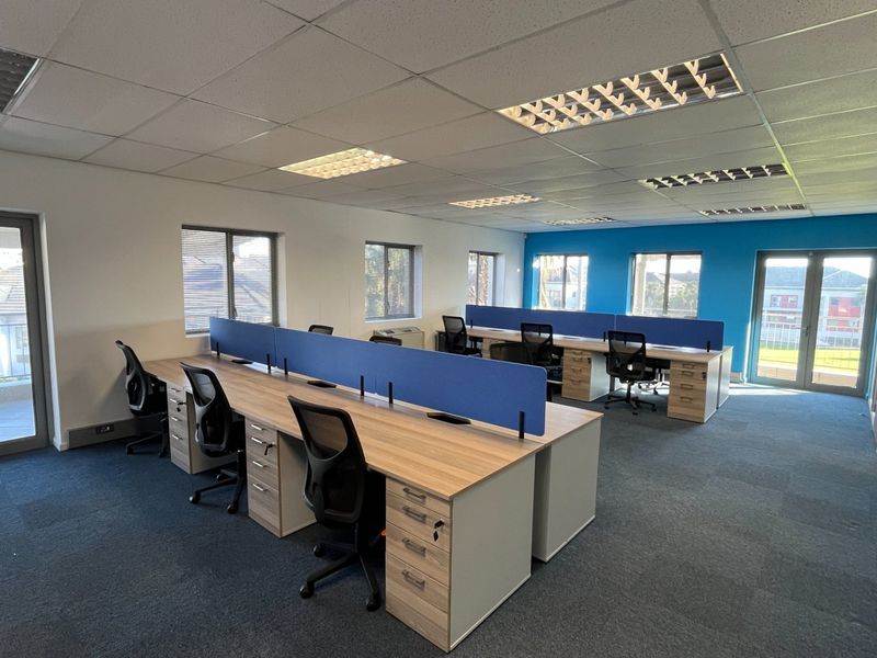 A modern, well-lit commercial office space featuring built-in desk workstations, carpeted flooring, and access to a balcony through double glass doors.