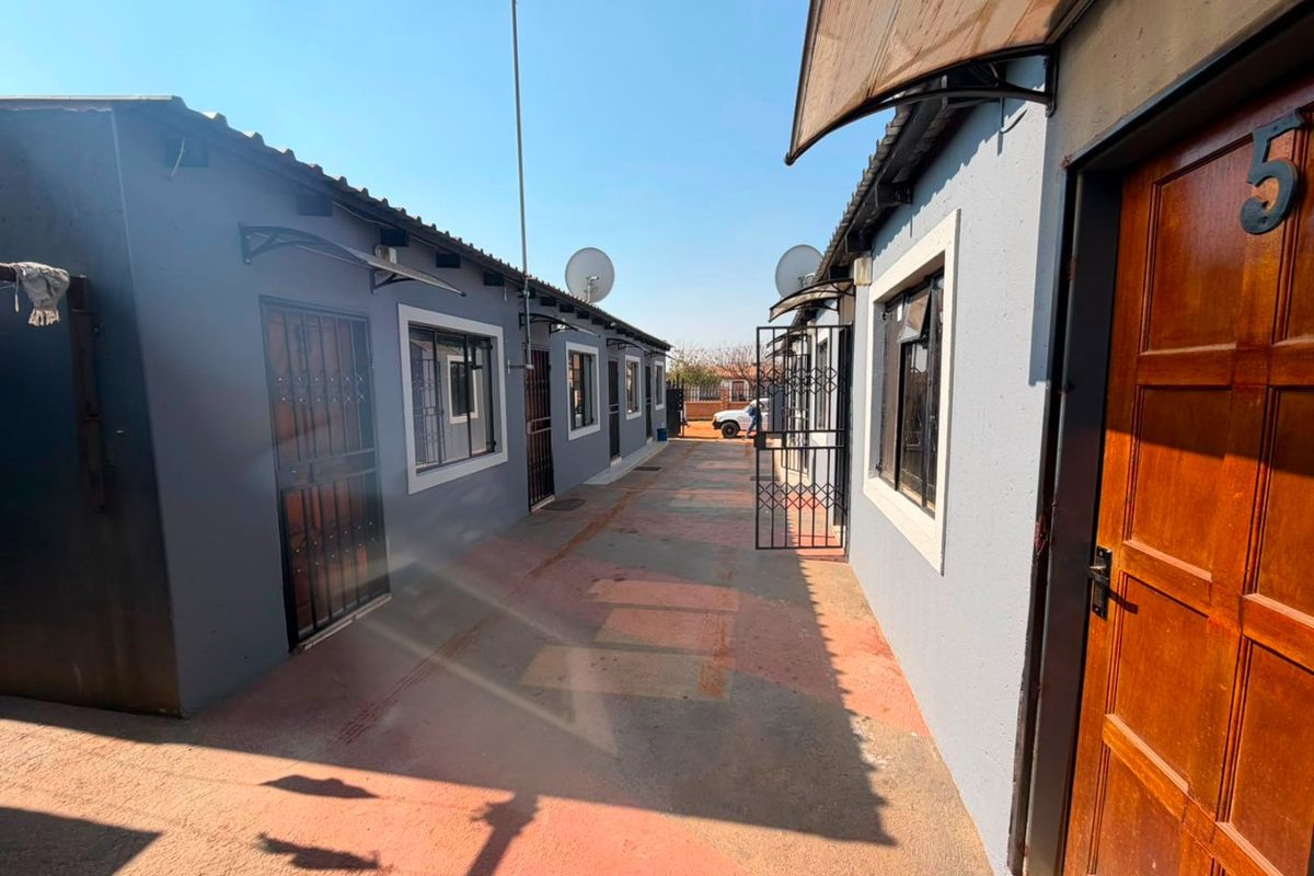 A narrow paved exterior walkway between two residential units featuring security gates, satellite dishes, and external doors.
