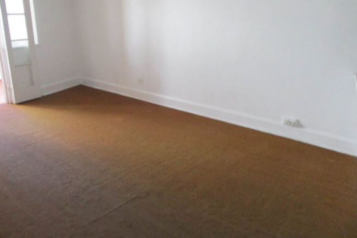 An empty living room featuring plain white walls, brown carpeting, and a classic multi-pane door leading to an adjacent space.