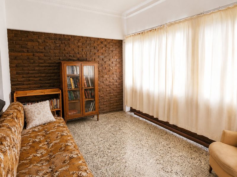 A living room featuring a textured exposed brick wall, classic terrazzo flooring, and large windows with natural light.