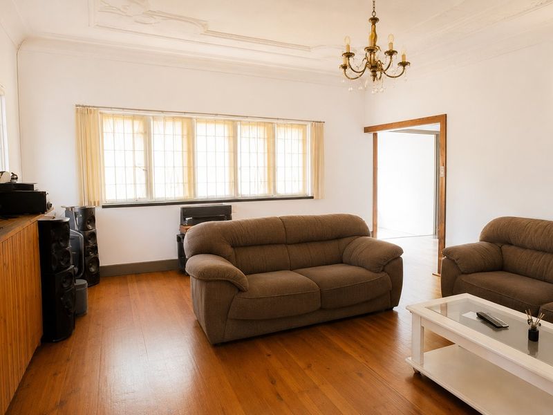 A spacious living room featuring polished hardwood flooring, ornate ceiling detailing, and a classic chandelier, offering a bright and inviting atmosphere.